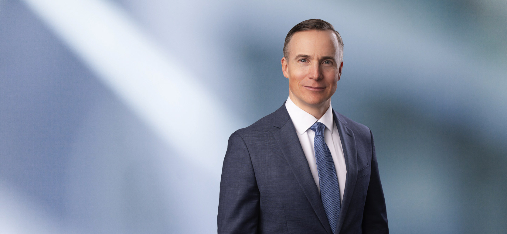 A man, Joel Farrar, in a blue suit, white shirt, and blue tie is looking at the camera, standing against a blurred blue and white background.