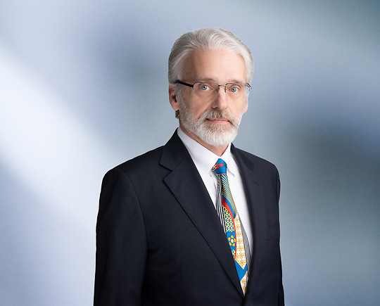 "A man, Michael Oliva, with short, dark brown hair, wearing a black blazer, is smiling at the camera, standing against a blurred blue and white background."