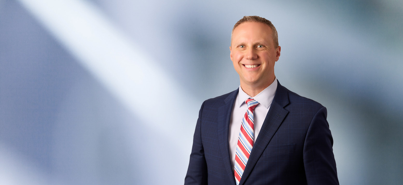 man, Todd Hoppe, with short light brown hair in a dark blazer, with a blurred blue and white background.