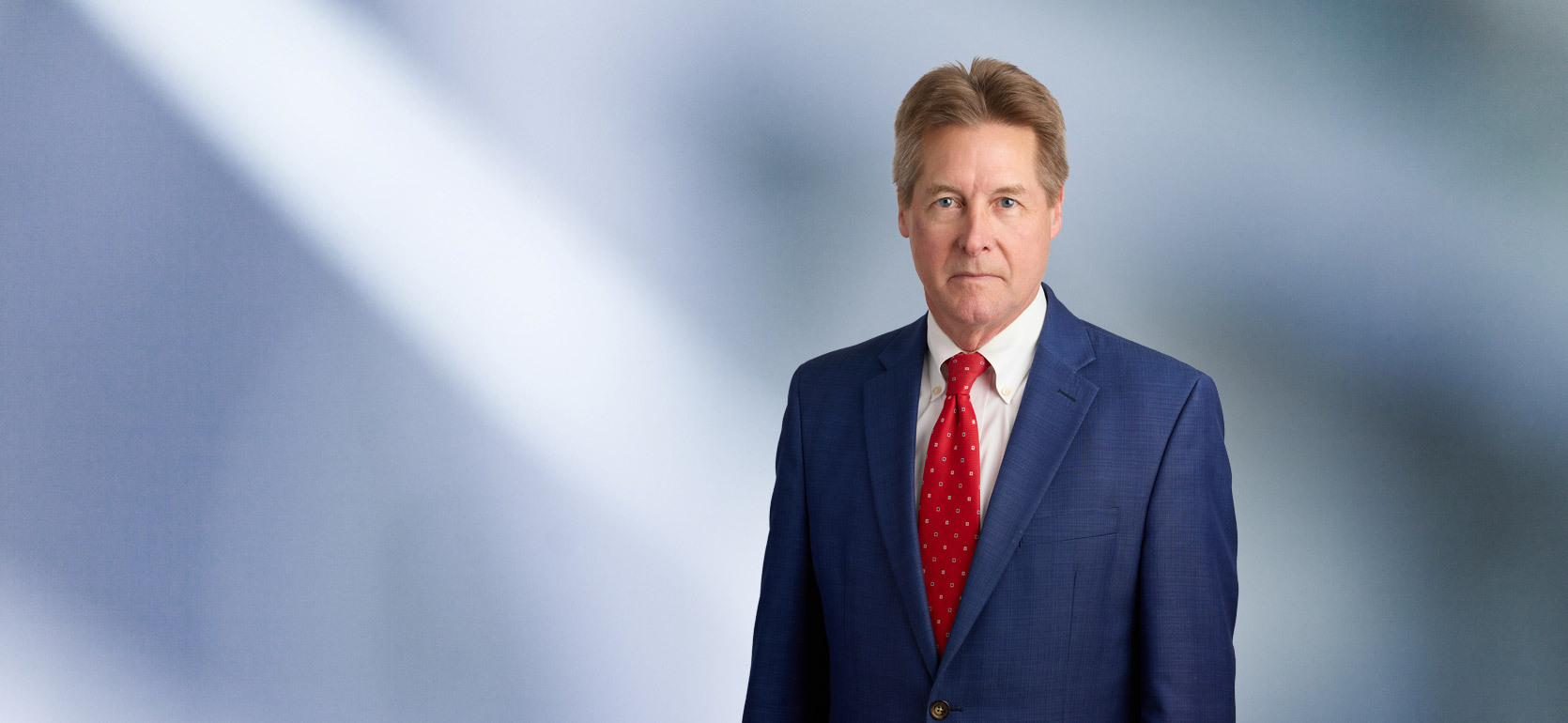 A man, James Doezema, in a dark suit, light shirt, and striped tie is smiling at the camera, standing against a blurred blue and white background.