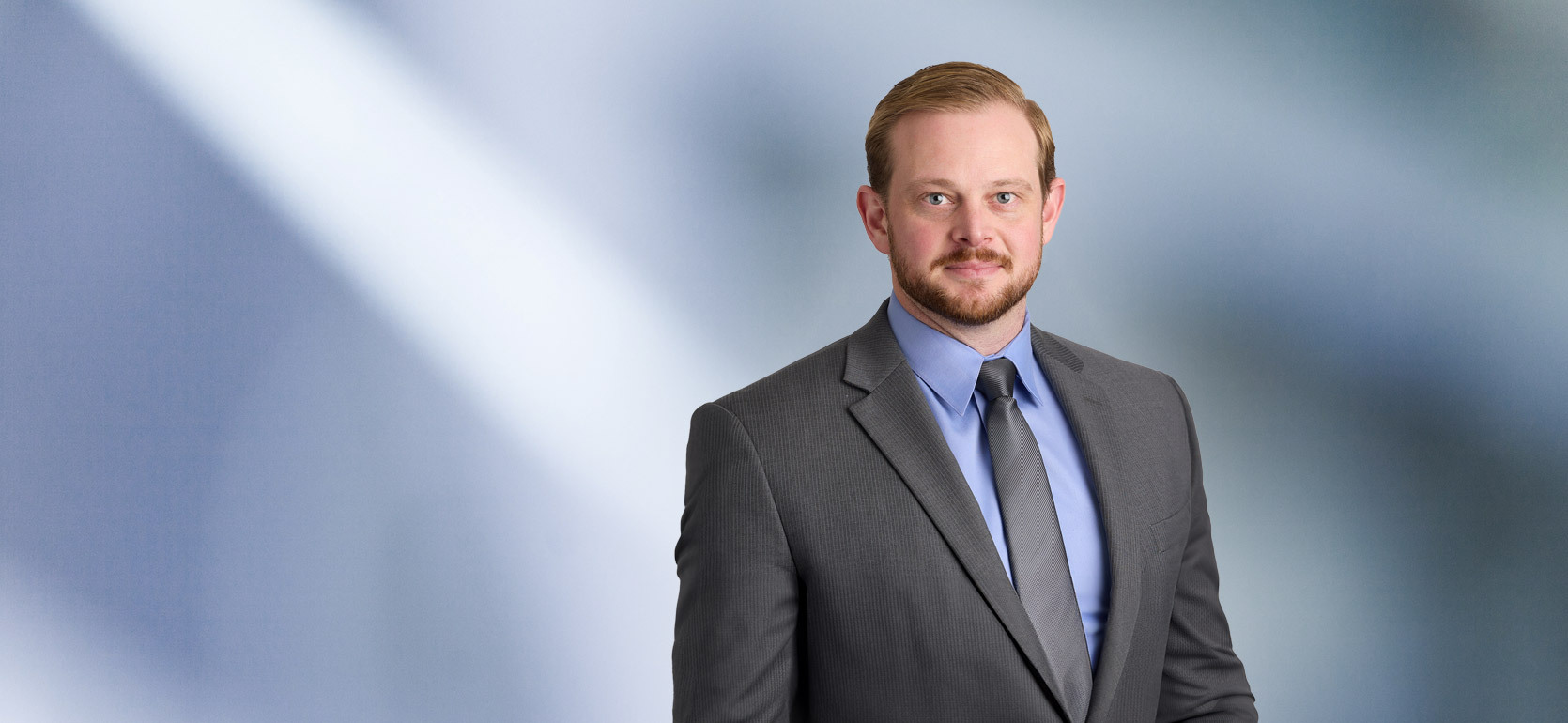 A man, Michael Zahrt, in a dark suit, light shirt, and striped tie is smiling at the camera, standing against a blurred blue and white background.