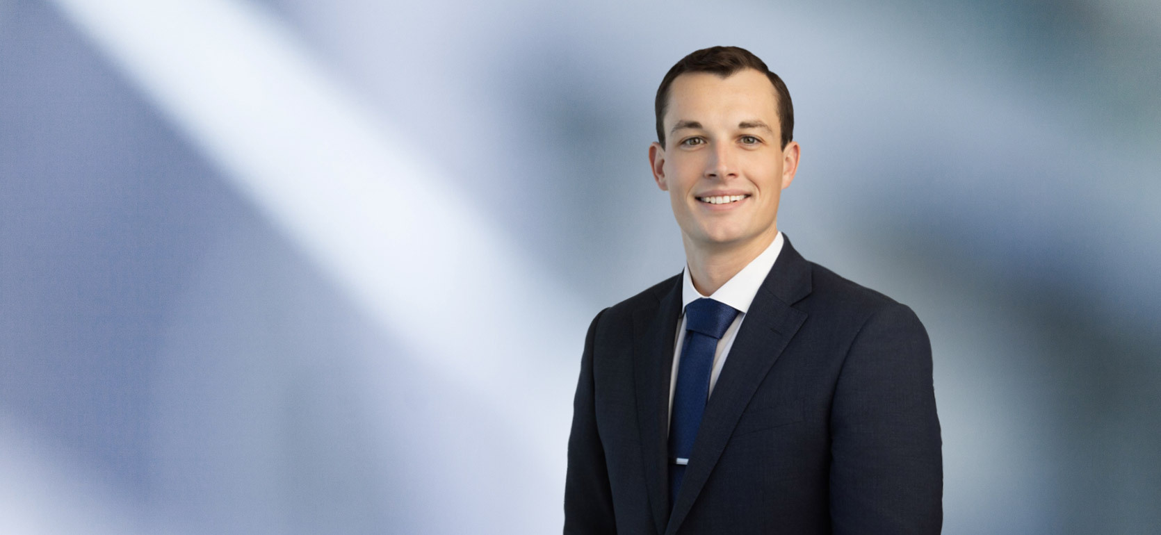 A man James Ryan, in a dark suit, white shirt, and blue tie is smiling at the camera, standing against a blurred blue and white background.