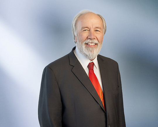 man, Dirk Beckwith, with a white beard wearing a dark suit, white shirt, and bright red tie, standing against a softly blurred blue and white background, smiling warmly at the camera.