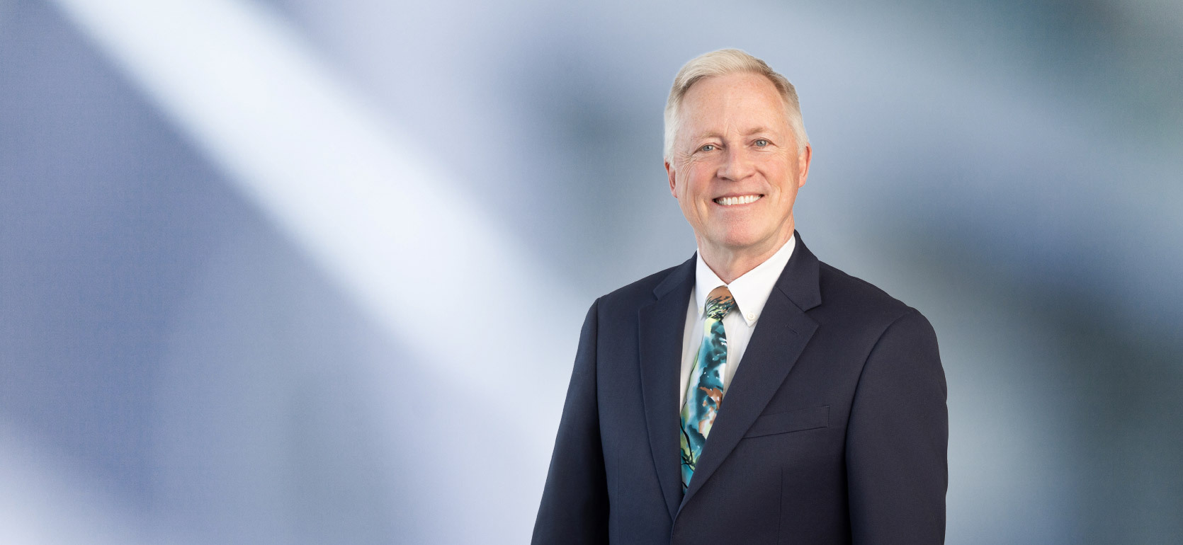 Professional man, Michael Liddane, with light hair wearing a dark suit, white shirt, and colorful patterned tie, standing against a softly blurred blue and white background