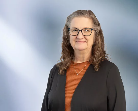A woman, Susan Paules, in a brown sweater and dark rimmed glasses is looking at the camera, standing against a blurred blue and white background.