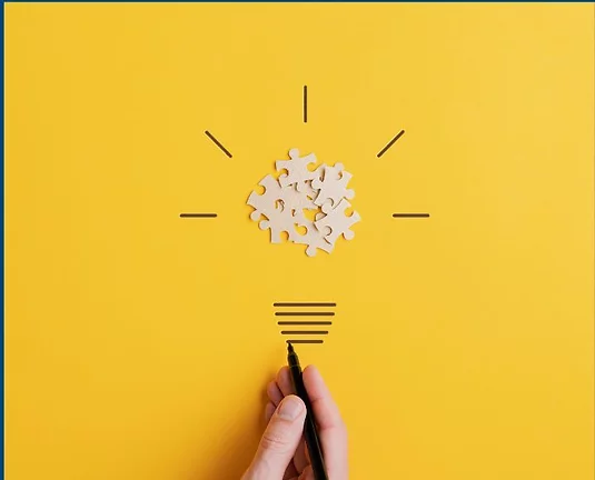 Paper puzzle pieces with hand drawn lines around to look like a lightbulb on a mustard yellow background.