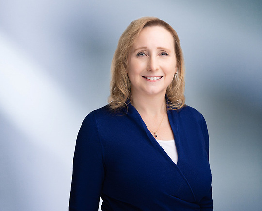 A woman, Ann Stevens, in a blue top and white blouse is smiling at the camera, standing against a blurred blue and white background.