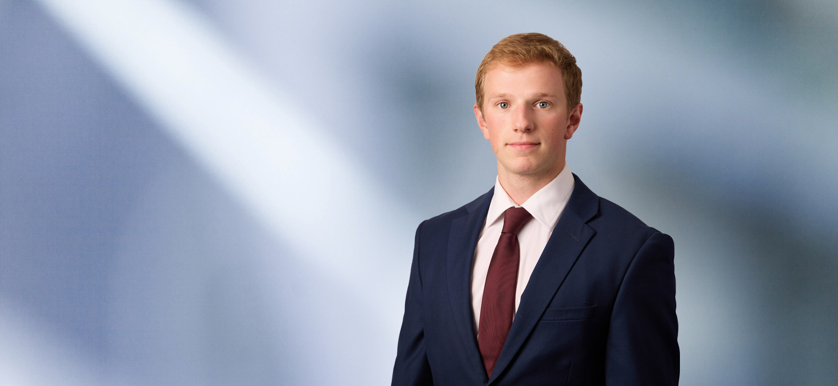A man, Charles Sarchet, in a dark suit, white shirt, and maroon tie is looking at the camera, standing against a blurred blue and white background.