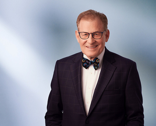 A man, Fredrick Dilley, in a dark checkered suit and a colorful bow tie is smiling at the camera, standing against a blurred blue and white background.
