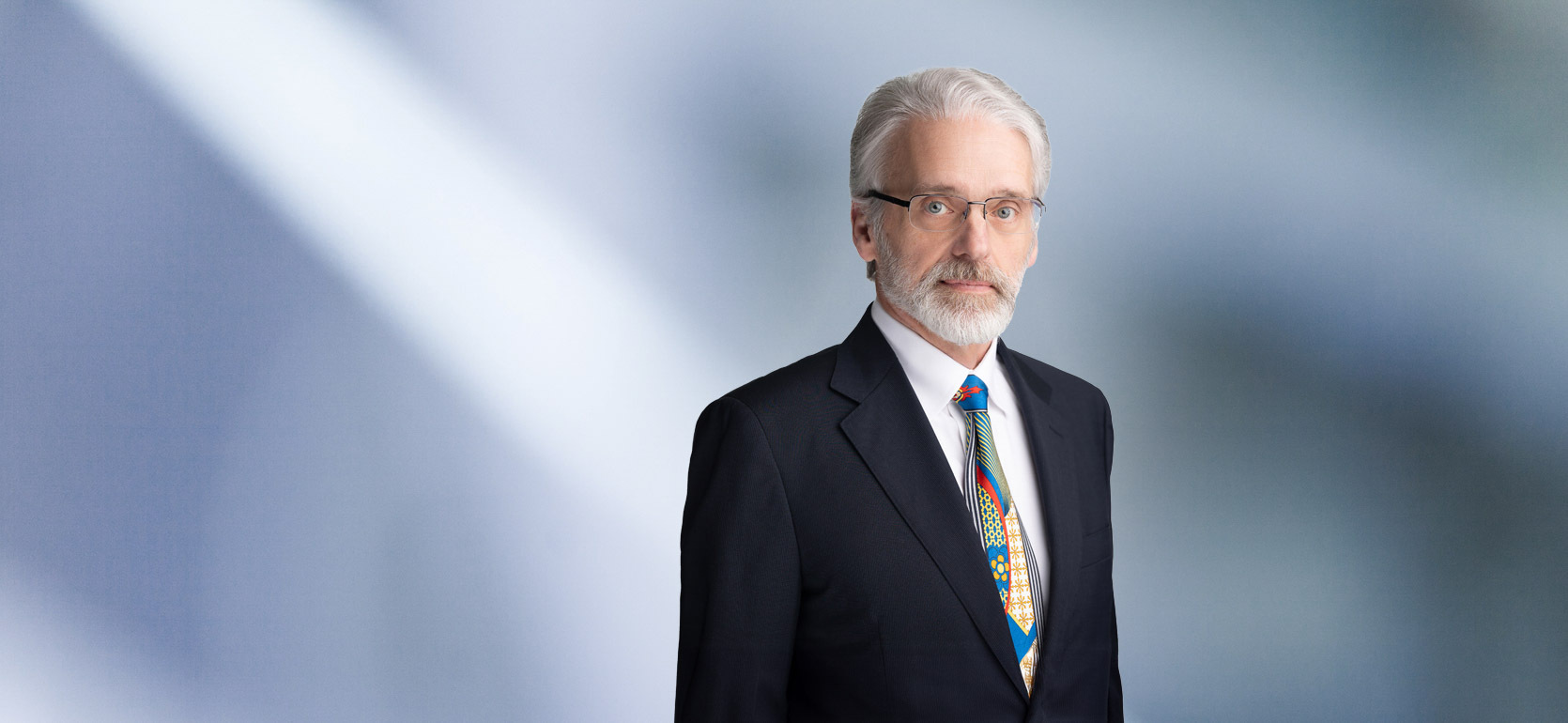 "A man, Michael Oliva, with short, dark brown hair, wearing a black blazer, is smiling at the camera, standing against a blurred blue and white background."