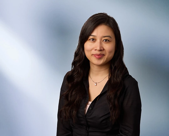 Portrait of a woman, Thailan Hang, with long black hair in a dark blazer, with a blurred blue and white background.
