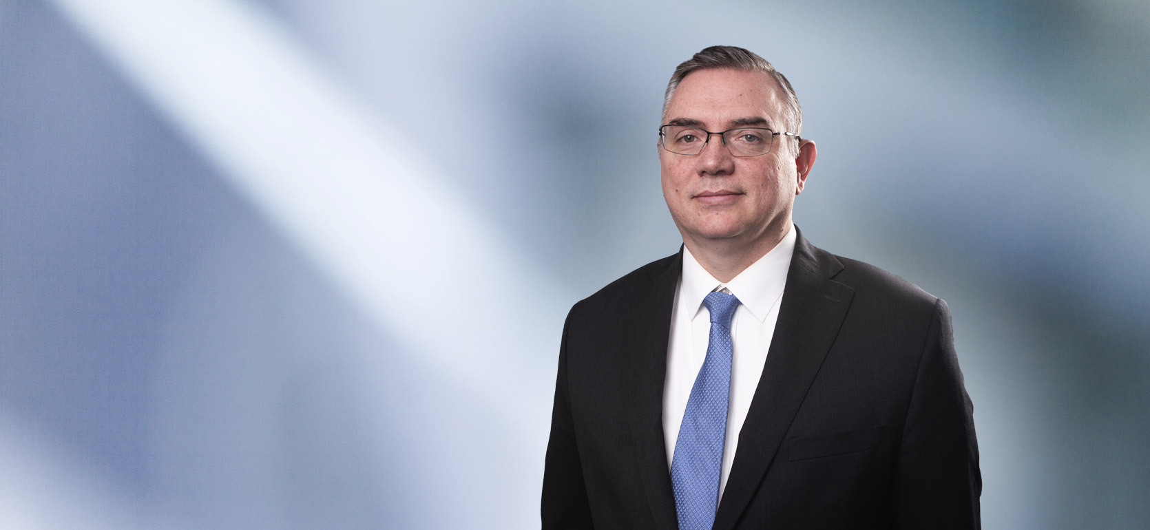 A man, Clifford Hammond, in a dark suit, white shirt, and light blue tie is looking at the camera, standing against a blurred blue and white background.