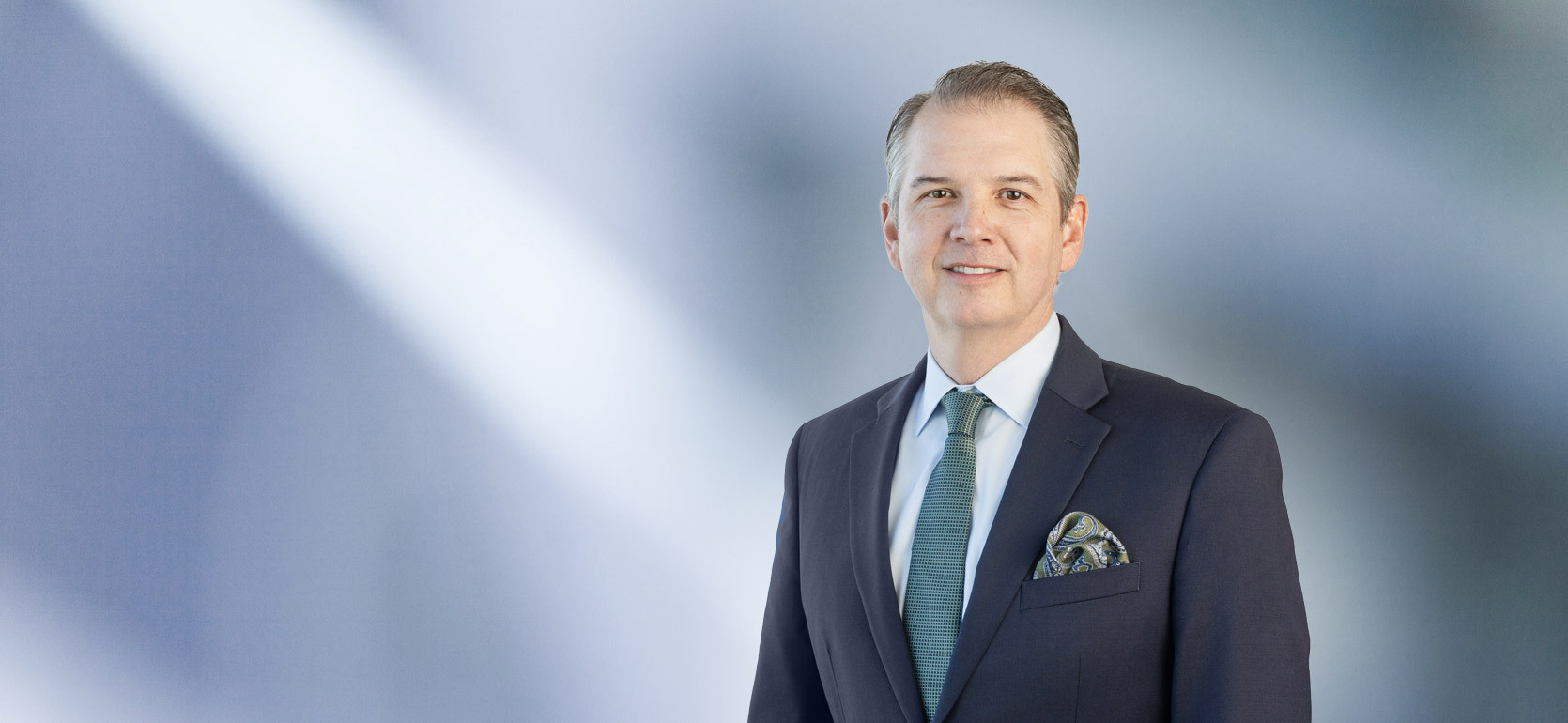 man, Matthew Fedor, wearing a dark suit, light blue shirt, green patterned tie, and matching pocket square, standing against a softly blurred blue and white background