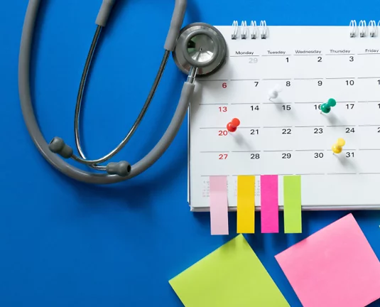 A stethoscope rests on a calendar with multicolored pushpins marking different dates. Bright sticky notes and page flags are placed at the bottom of the calendar, all set against a vibrant blue background—suggesting healthcare appointment planning or a me