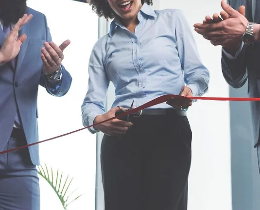 Businesswoman cutting a red ribbon at a ribbon-cutting ceremony, surrounded by colleagues clapping and celebrating the event.