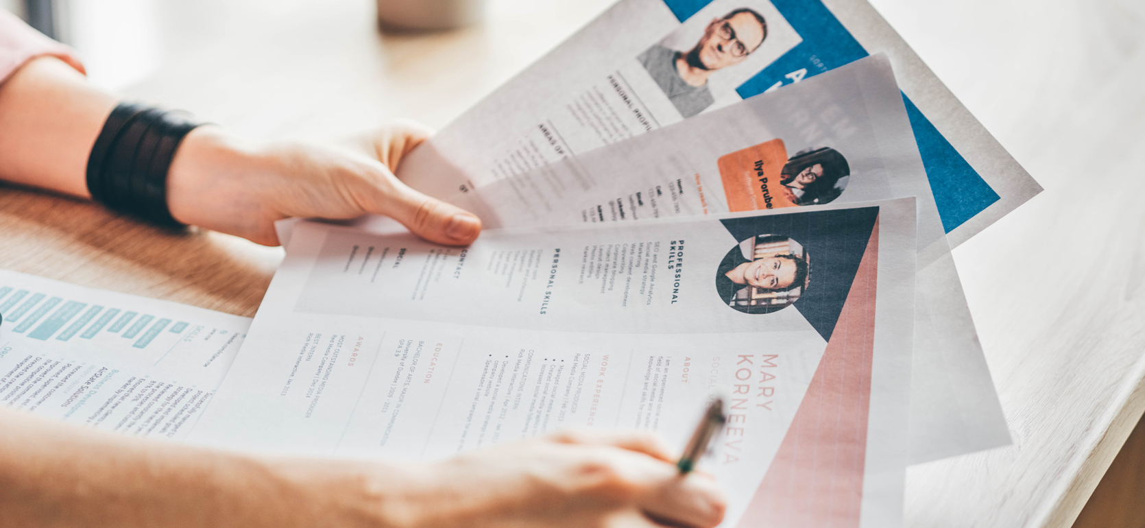 One hand holding three paper resumes at desk while other hand holds pen writing notes