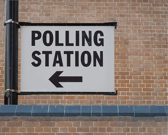 A white sign with the words "Polling Station" and an arrow pointing to right in black tied to a pole with a brick wall in the background.