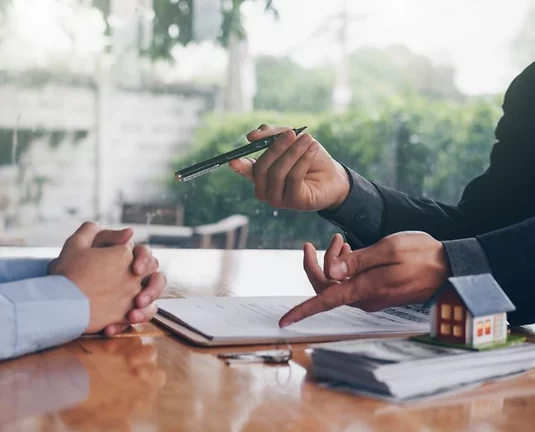 Two people negotiating over a contract with a small house model and house keys on the table.