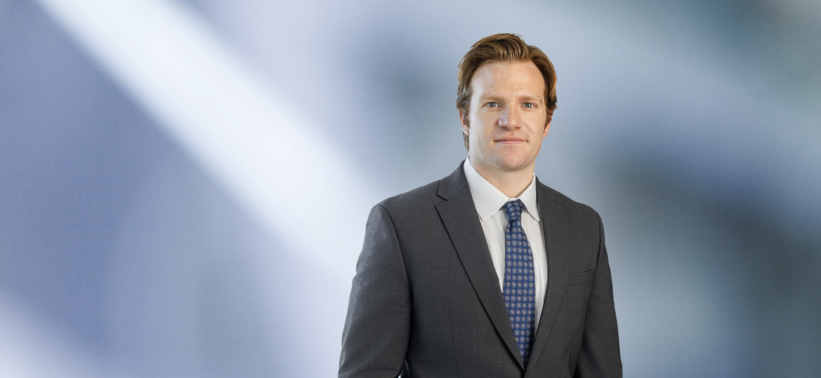 A man, Anthony Dalimonte, in a gray suit, blue shirt, and blue striped tie is smiling at the camera, standing against a blurred blue and white background.