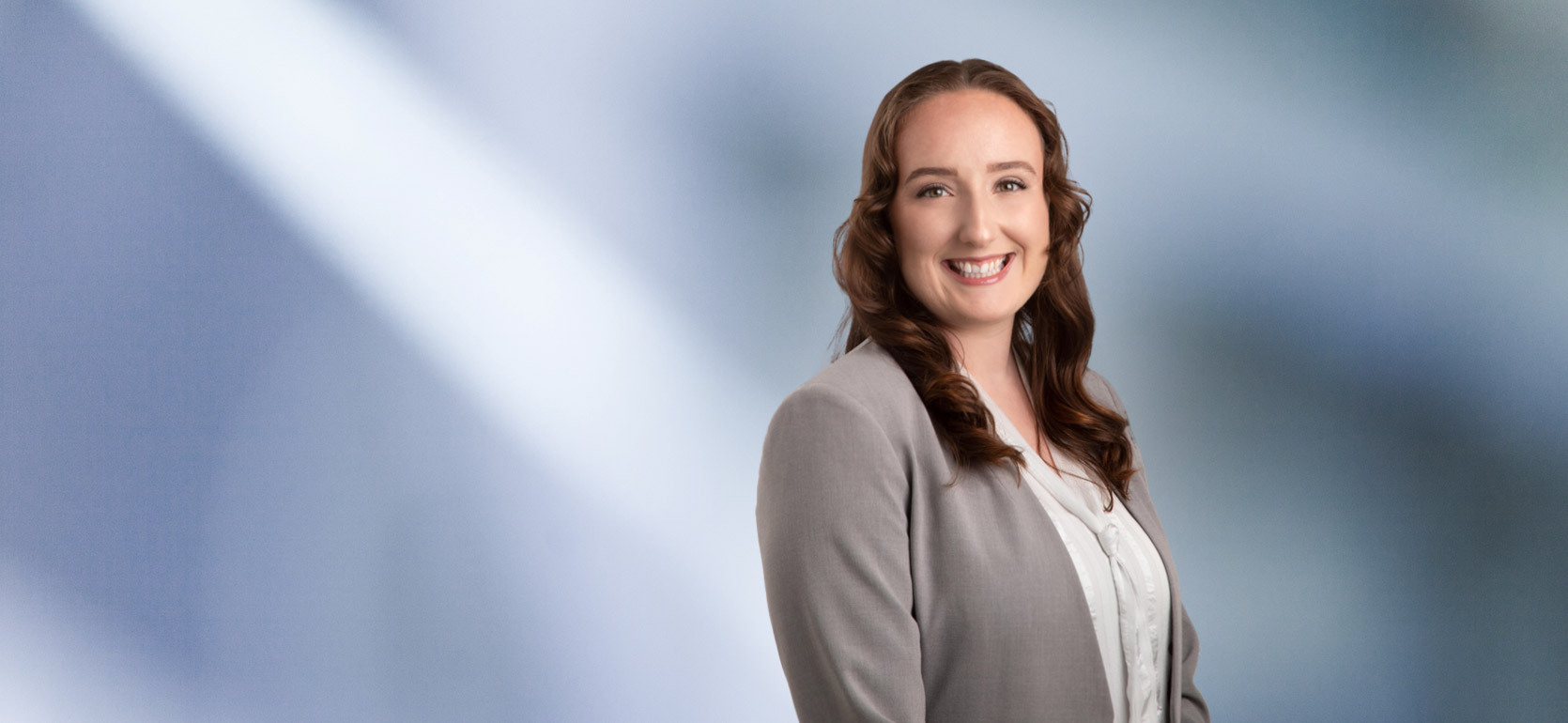 woman, Destiny Hughes, wearing a light gray blazer and white blouse, standing against a softly blurred blue and white background, smiling confidently at the camera