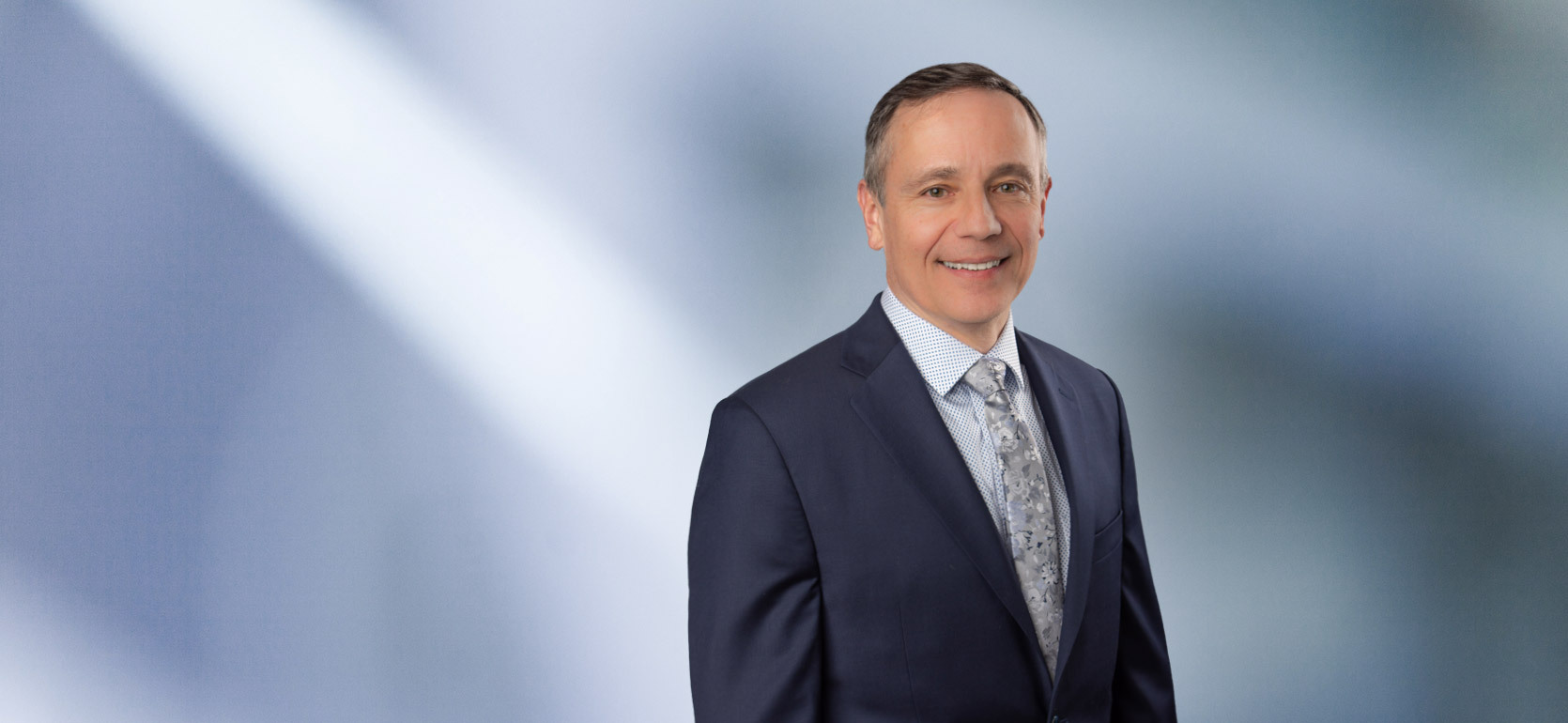 “A man, Matt Hrebec, with short, light brown hair, wearing a grey blazer and white collared shirt, is smiling at the camera, standing against a blurred blue and white background.”