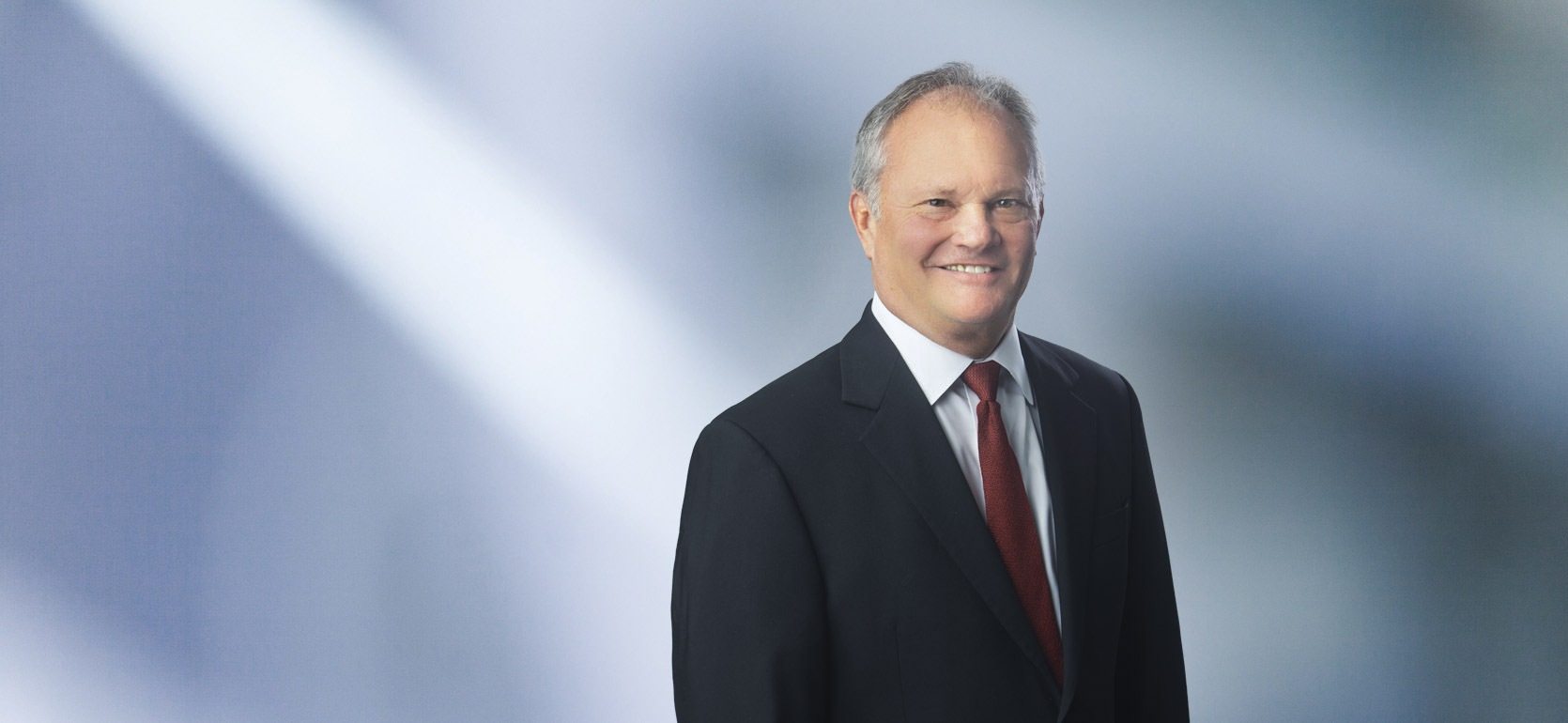 A man, Brian Goodenough, in a dark suit, white shirt, and red tie is smiling at the camera, standing against a blurred blue and white background.