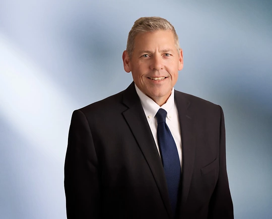 "A man, Michael Homier, with short, light brown hair, wearing a dark blazer, is smiling at the camera, standing against a blurred blue and white background."