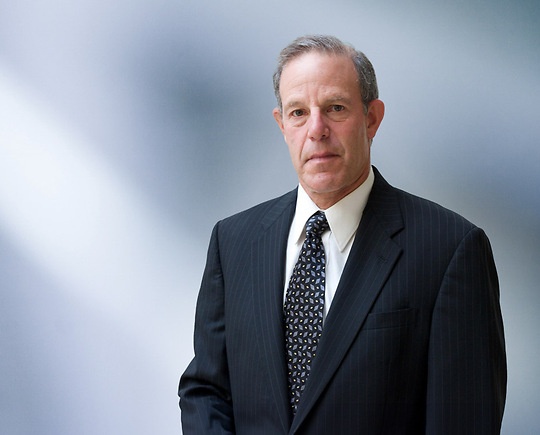 Man, Scott Mandel, wearing a dark blazer, standing against a blurred blue and white background.