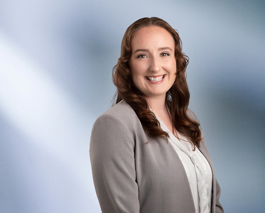 woman, Destiny Hughes, wearing a light gray blazer and white blouse, standing against a softly blurred blue and white background, smiling confidently at the camera