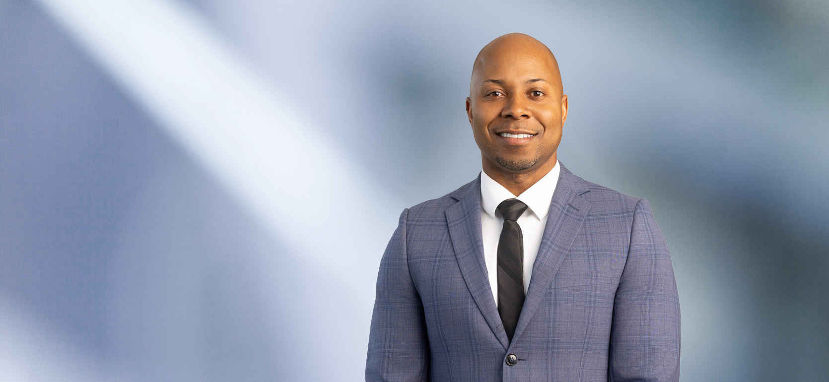 A bald African American man, Ray Littleton, wearing a blue tweed pattern sport coat with a white undershirt and black striped tie is smiling at the camera, standing against a blurred blue and white background.
