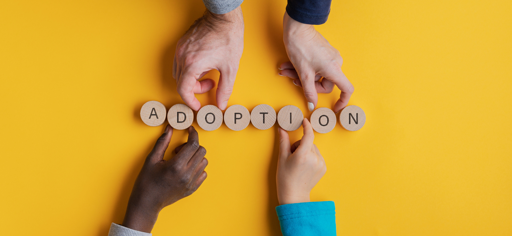 Four hands of multiple ages holding block letters that spell adoption over yellow background