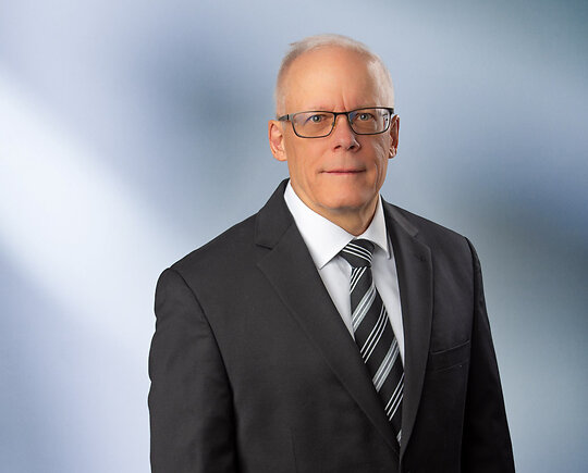 A man, Brent Titus, in a gray suit, light blue shirt, and red patterned tie is looking at the camera, standing against a blurred blue and white background.