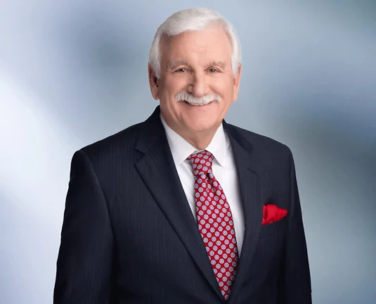 Older man, Mike Kluck smiles at camera in front of grey background wearing a black pinstripe jacket and red dotted tie