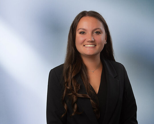 A young dark-haired woman, Rachel Conner, wearing a black sport coat and black blouse, smiles at camera in front of blurred blue-white background