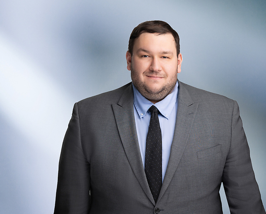 A man, Brandon Schumacher, in a gray suit, light blue shirt, and black tie is smiling at the camera, standing against a blurred blue and white background.