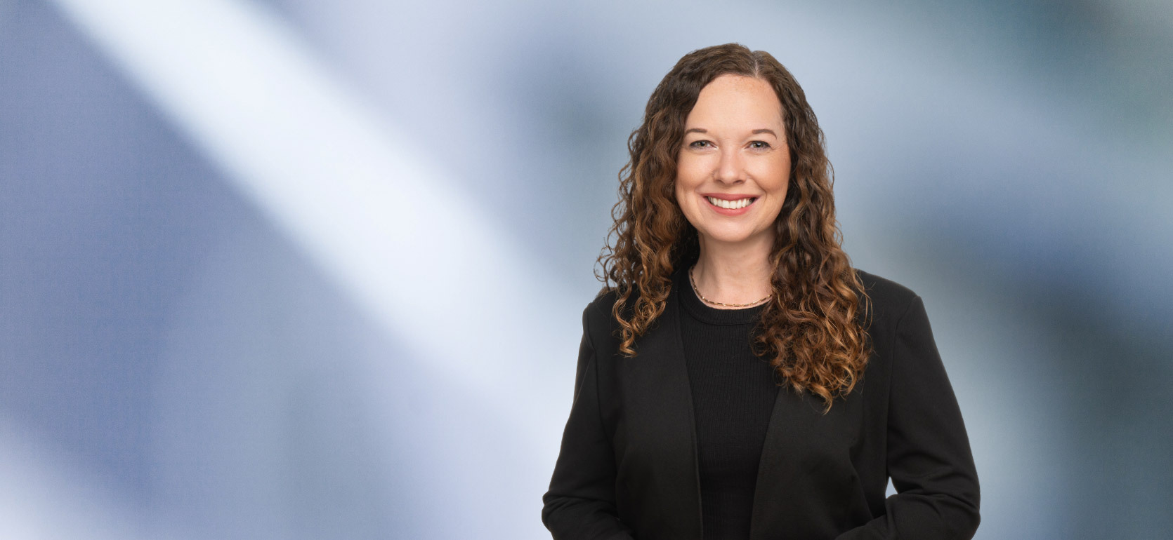 A woman, Laura Genovich, with curly brown hair, wearing a black blazer and black top, is smiling at the camera, standing against a blurred blue and white background.