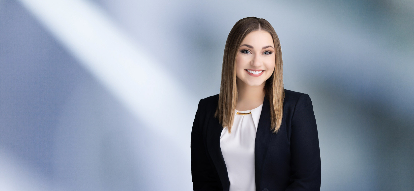 A woman, Lindsey Mead, with straight, light brown hair, wearing a navy blazer and white top with a gold accent, is smiling at the camera, standing against a blurred blue and white background.