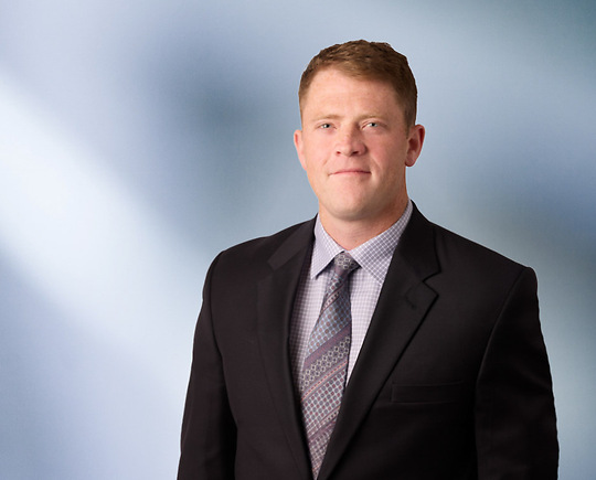 Man, Nicholas Stock, with short dark brown hair wearing a blue blazer, smiling against a blurred white and blue background.
