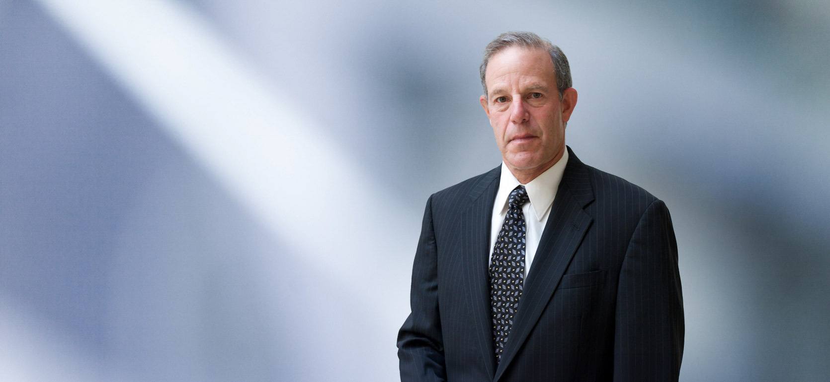 Man, Scott Mandel, wearing a dark blazer, standing against a blurred blue and white background.