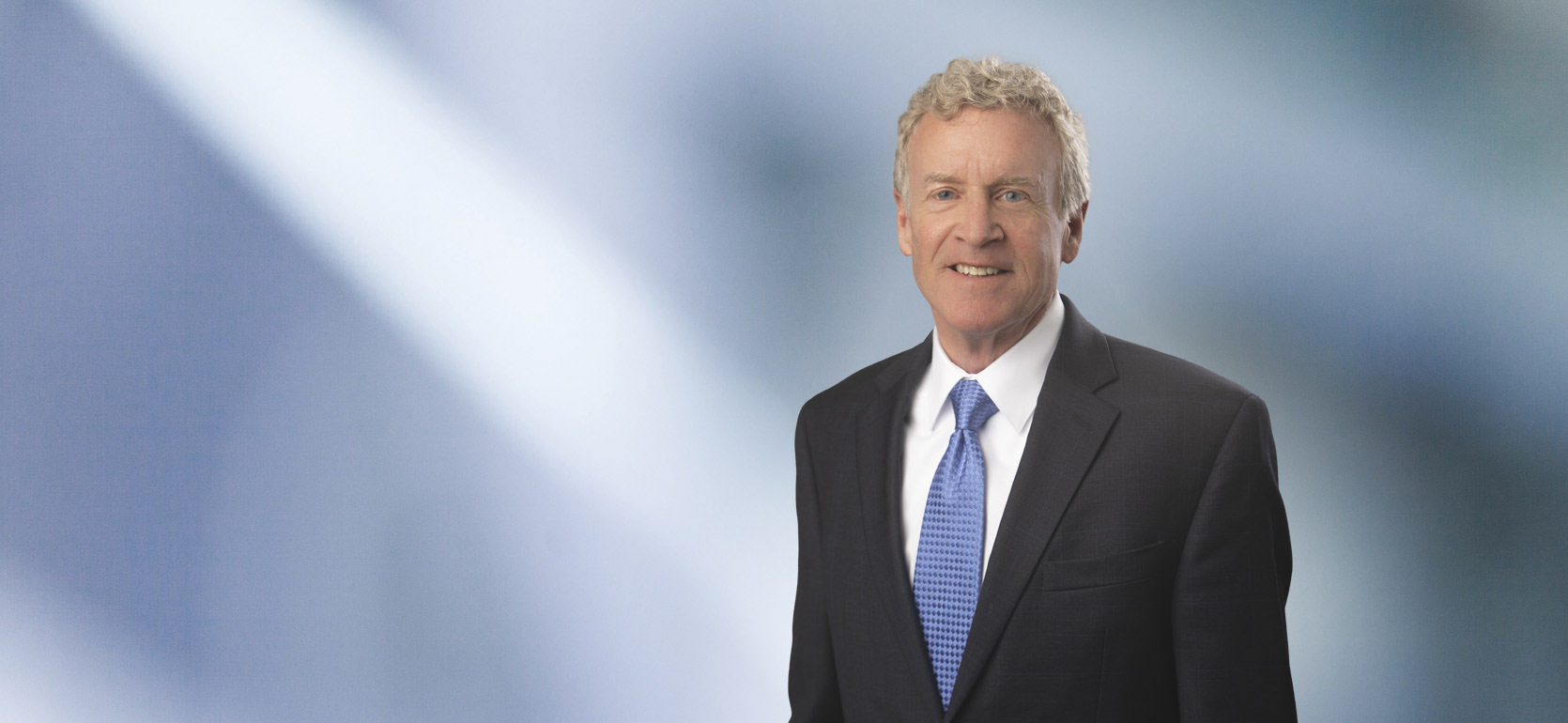 Portrait of a man, Thomas Meagher, with short brown hair in a dark blazer, with a blurred blue and white background.