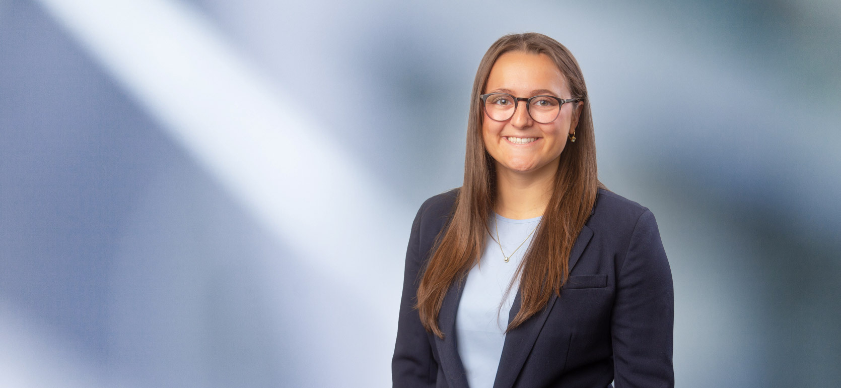 A young dark-haired woman with glasses, Olivia Reid, wearing a black sport coat and light blue blouse, smiles at camera in front of blurred blue-white background