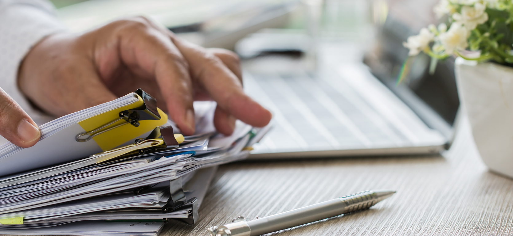 Hand looking through packets of paper held together on desk with pen and laptop