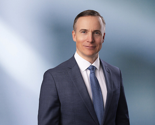 A man, Joel Farrar, in a blue suit, white shirt, and blue tie is looking at the camera, standing against a blurred blue and white background.