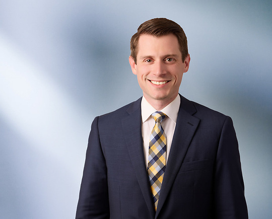 Portrait of a man, Thomas Dillon, with short brown hair in a dark blazer, with a blurred blue and white background.