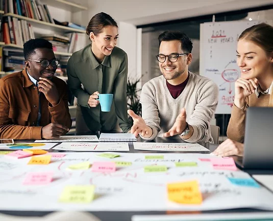 Four diverse coworkers collaborating at a table covered in colorful sticky notes and planning charts. One person is talking and gesturing, while others listen and smile in a creative office setting.