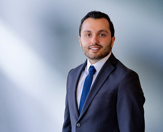 A young man, Badri Yono, wearing a blue-gray blazer, white shirt and solid blue time, smiles at camera in front of blurred blue-white background