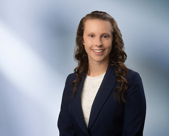 A young woman, Allison Shudark, wearing a black sport coat and white lace blouse, smiles at camera in front of blurred blue-white background