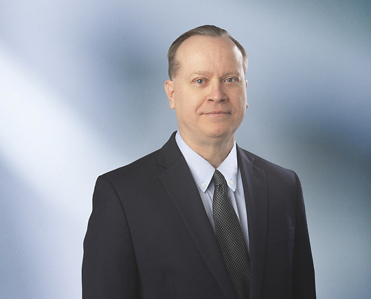 A man, Doug Mielock, in a black suit, light blue shirt, and dark patterned tie is looking at the camera, standing against a blurred blue and white background.