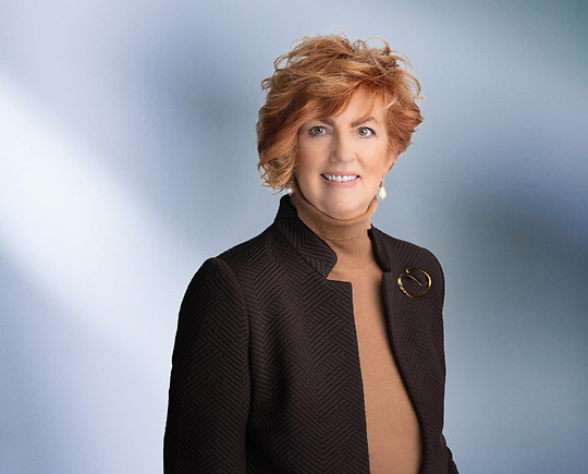 A woman, Kelly Lucas, in a brown textured blazer over a tan turtleneck, accessorized with pearl earrings and a brooch, is smiling at the camera, standing against a blurred blue and white background.
