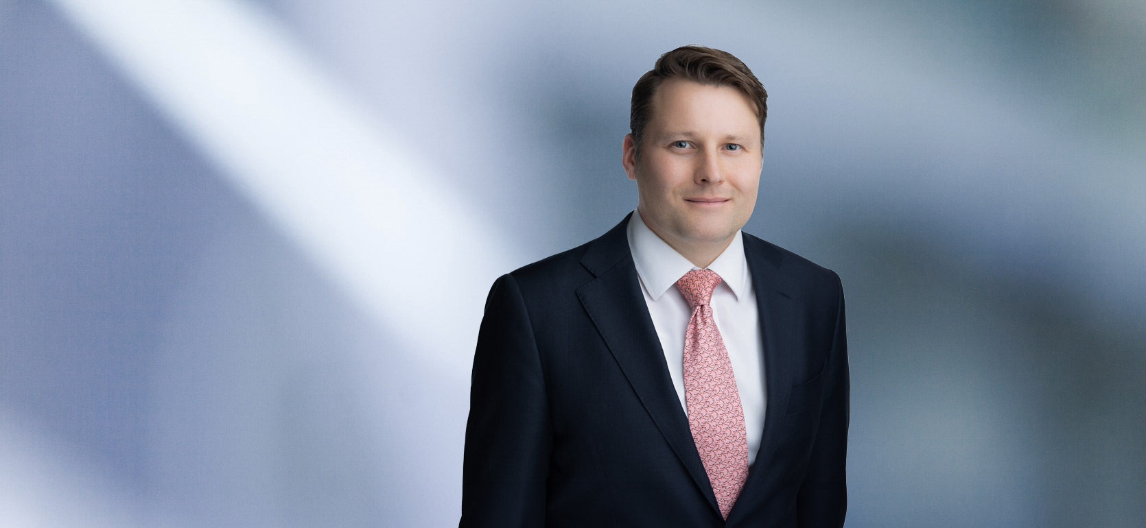 A man, Benjamin Williams, in a dark suit, white shirt, and light pink patterned tie is smiling at the camera, standing against a blurred blue and white background.